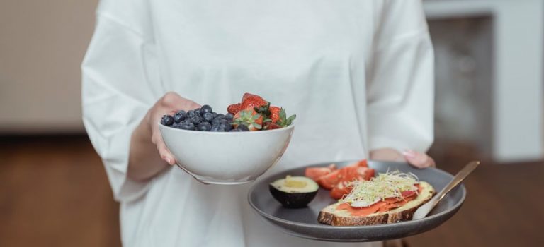 Person holding a bowl of fresh berries and a plate with avocado toast, displaying a healthy breakfast spread.